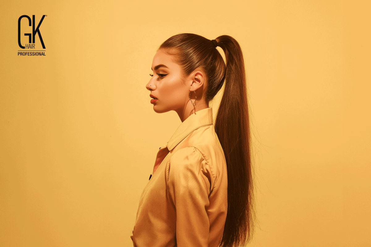 A person with long, straight hair tied in a ponytail, wearing a yellow blouse, set against a warm yellow background.